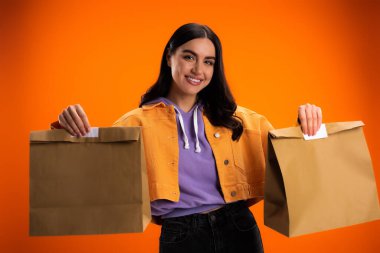 Positive woman holding paper bags and looking at camera isolated on orange