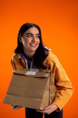 Young brunette woman holding paper bags and looking away isolated on orange