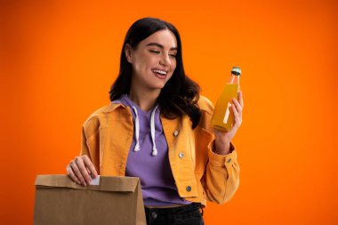 Cheerful woman holding paper bag and bottle of juice isolated on orange