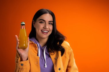 Cheerful brunette woman holding bottle with juice isolated on orange