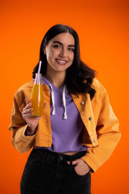 Smiling young woman holding bottle with juice and drinking straw isolated on orange
