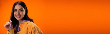 Pretty brunette woman holding juice with dinking straw isolated on orange, banner 