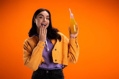 Excited woman looking at camera while holding bottle with juice isolated on orange