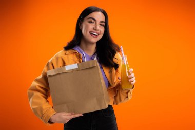 Cheerful young woman holding paper bag and juice in glass bottle isolated on orange