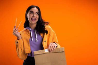 Joyful brunette woman holding chopsticks and paper bag isolated on orange