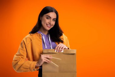 Smiling brunette woman holding bamboo chopsticks and paper bag isolated on orange