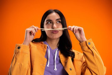 Young woman holding bamboo chopsticks near mouth and pouting lips isolated on orange