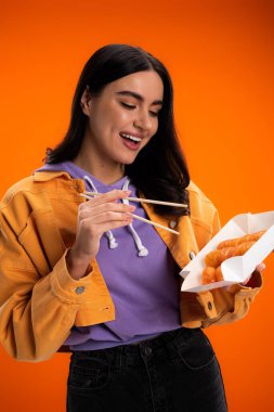 Positive brunette woman holding chopsticks and sushi in package isolated on orange
