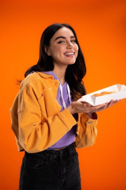 Positive woman holding takeaway sushi and smiling at camera isolated on orange