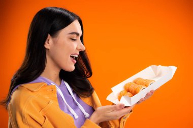 Side view of cheerful woman looking at takeaway sushi isolated on orange