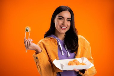 Smiling brunette woman holding sushi and looking at camera isolated on orange