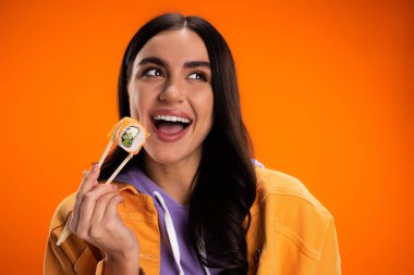 Portrait of excited woman holding sushi in chopsticks isolated on orange