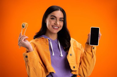 Positive young woman holding sushi in chopsticks and smartphone with blank screen isolated on orange