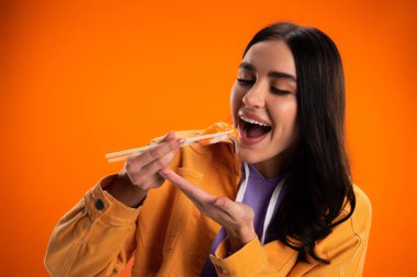 brunette woman in stylish jacket eating delicious sashimi isolated on orange