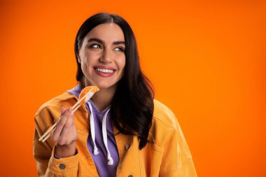 smiling woman in trendy jacket holding chopsticks with fresh sashimi isolated on orange clipart