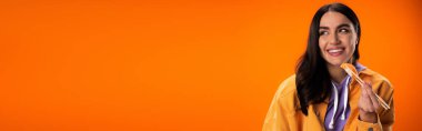 pleased brunette woman holding sashimi with chopsticks and looking away isolated on orange, banner