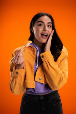 amazed woman holding delicious sashimi while touching face and looking at camera isolated on orange