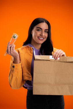 happy brunette woman with paper bag and sushi roll looking at camera isolated on orange