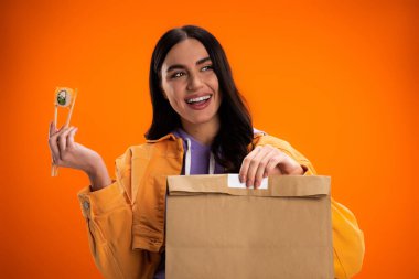 joyful woman holding paper bag and chopsticks with sushi roll while looking away isolated on orange