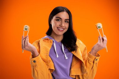 young brunette woman smiling at camera while holding chopsticks with sushi rolls isolated on orange