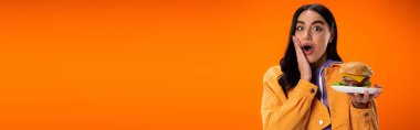 astonished woman with delicious burger touching face and looking at camera isolated on orange, banner
