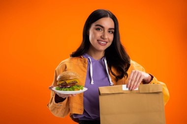 happy brunette woman holding paper bag and delicious burger isolated on orange