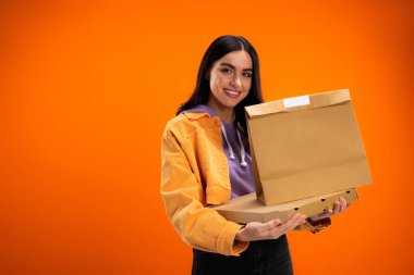 brunette woman with pizza box and paper bag smiling at camera isolated on orange
