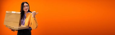 smiling woman with food packages pointing with thumb and looking away isolated on orange, banner
