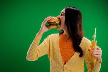 brunette woman holding bottle of fresh lemonade and eating tasty burger isolated on green