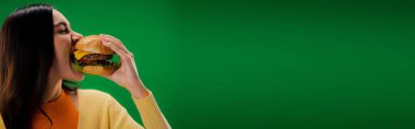 young and hungry woman biting tasty burger isolated on green, banner
