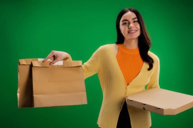 smiling woman holding craft paper bags and pizza box while looking at camera isolated on green