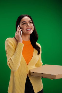 happy young woman calling on smartphone while standing with pizza box isolated on green