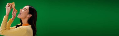 side view of young and hungry woman eating tasty pizza isolated on green, banner
