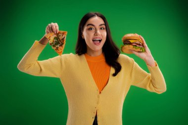 young amazed woman with piece of pizza and tasty burger looking at camera isolated on green