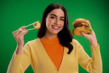 cheerful woman holding tasty burger and fresh sushi while looking at camera isolated on green