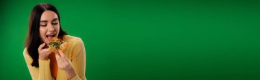young brunette woman in yellow jumper eating tasty pizza isolated on green, banner