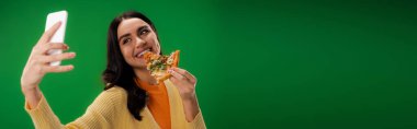 smiling brunette woman taking selfie on cellphone while eating tasty pizza isolated on green, banner clipart
