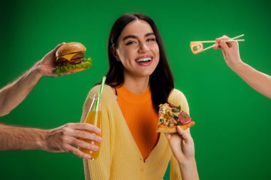happy young woman looking at camera near people proposing different food isolated on green
