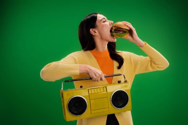 young and hungry woman with boombox eating tasty burger isolated on green