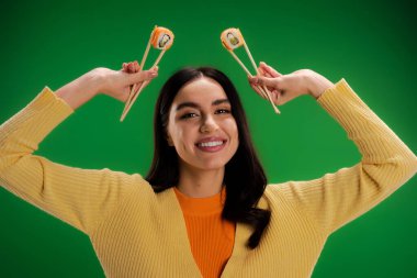 cheerful woman in yellow jumper holding fresh sushi rolls with chopsticks isolated on green