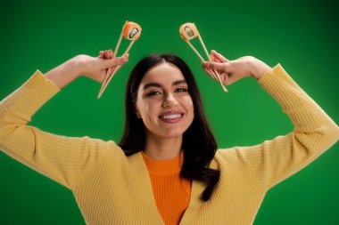 cheerful woman in yellow jumper holding fresh sushi rolls with chopsticks isolated on green clipart