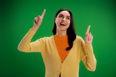 excited woman in yellow jumper looking up and pointing with fingers isolated on green clipart