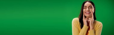 young brunette woman with closed eyes touching cheeks and smiling isolated on green, banner