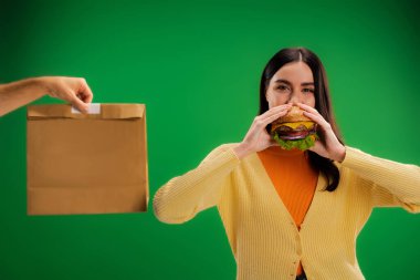 hungry woman eating delicious burger near man with food package isolated on green