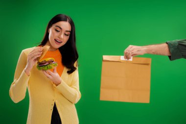 pleased woman looking at tasty burger near delivery man with paper bag isolated on green