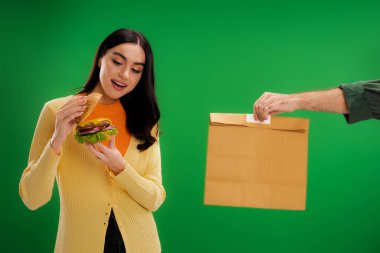 pleased woman looking at tasty burger near delivery man with paper bag isolated on green clipart