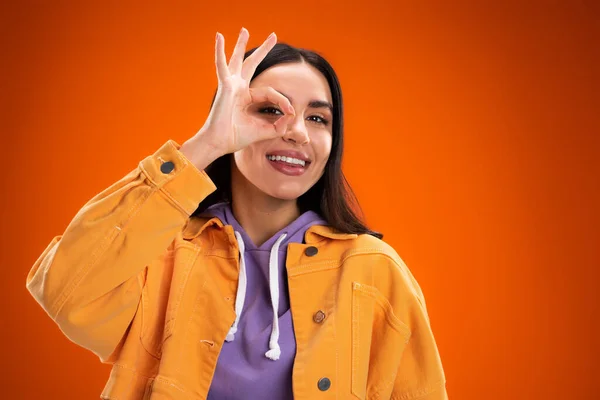 Cheerful woman in hoodie and jacket showing okay gesture isolated on orange