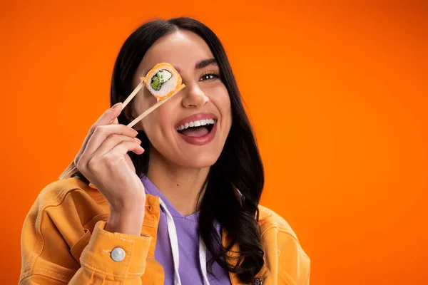 stock image Positive young woman holding sushi in chopsticks near eye isolated on orange