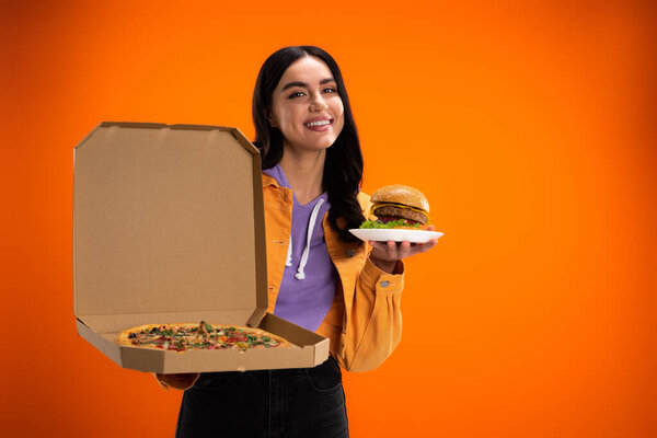happy young woman with delicious pizza and burger looking at camera isolated on orange