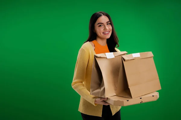 stock image happy brunette woman holding paper bags and pizza box while looking at camera isolated on green